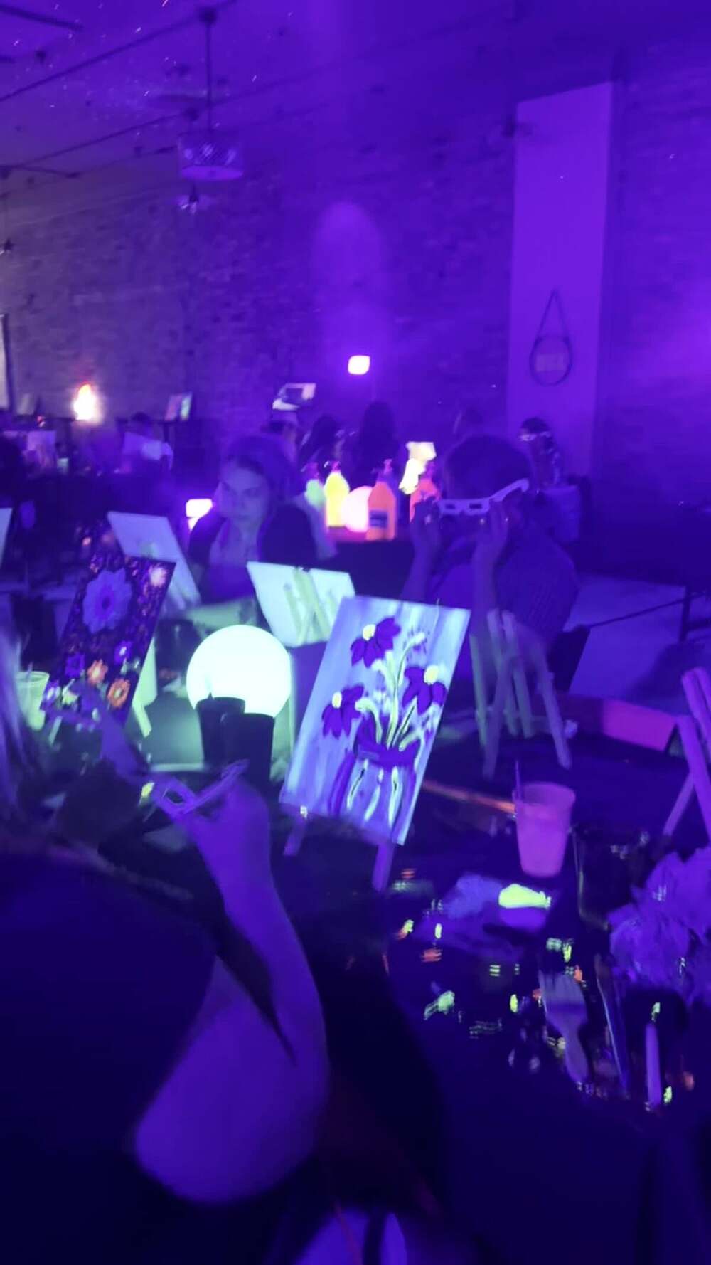 Cups of fluorescent UV paint glowing on a table beside a canvas
